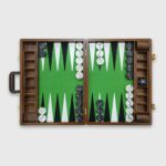 A tournament-sized Monte Carlo backgammon board with green and brown playing surfaces, accompanied by a set of black and white checkers, a doubling cube, and dice cups, with the board's case partially visible.